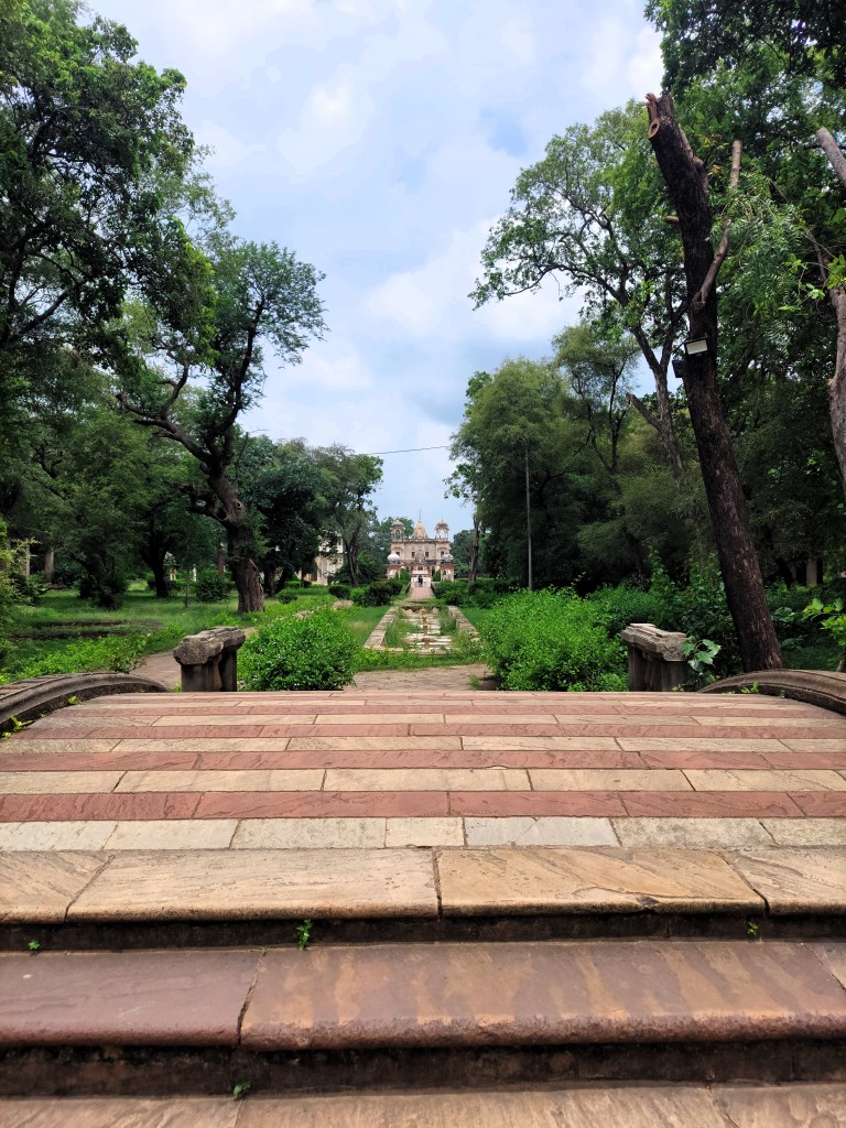 A small foot overbridge leads to a sandstone cenotaph, flanked by gardens on both sides.